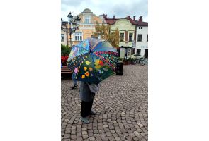 Osoba na rynku trzyma kolorowy parasol z wielobarwnymi wzorami, w tle kamienice.