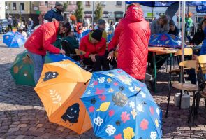 Grupa osób na rynku maluje kolorowe wzory na parasolach. Na pierwszym planie widać pomarańczowy i niebieski parasol ozdobione rysunkami.